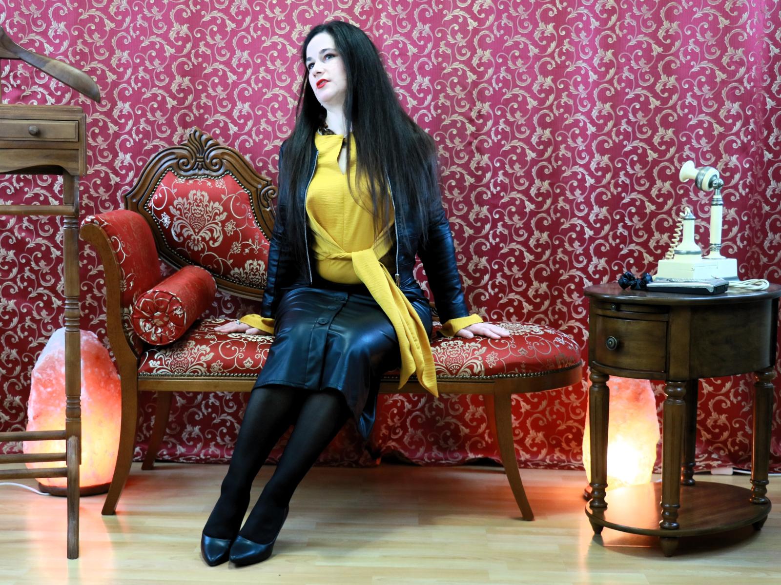 AmanelWhite with long black hair, red lipstick, yellow top, black leather skirt, black stockings, and black heels, sitting on a red and gold patterned sofa, wooden side table.
