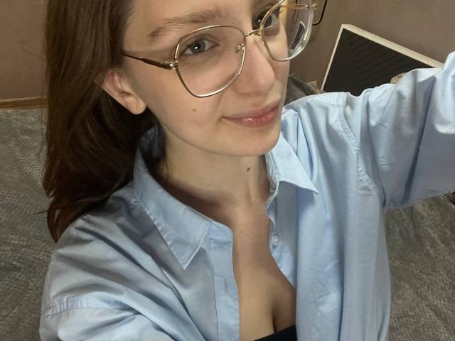 StaceyAverbach with glasses, light blue shirt, showing a bit of black bra, small tits, smiling, brown hair, light skin, standing, taking selfie, carpeted floor, warm lighting.