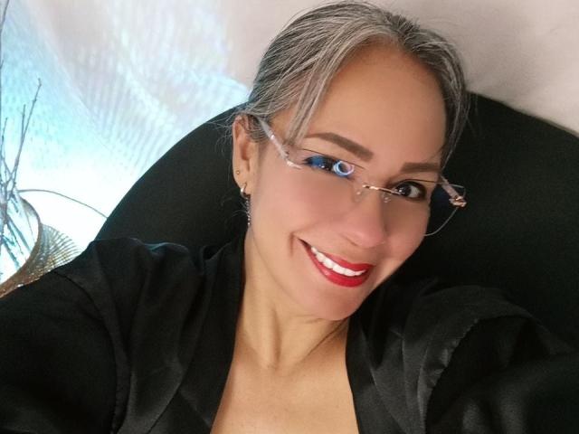 LailaCastillo with gray hair, blue eyeshadow, red lipstick, black satin robe, glasses, smiling, fair skin, black chair, white wall, and a lamp with a fan design in the background.