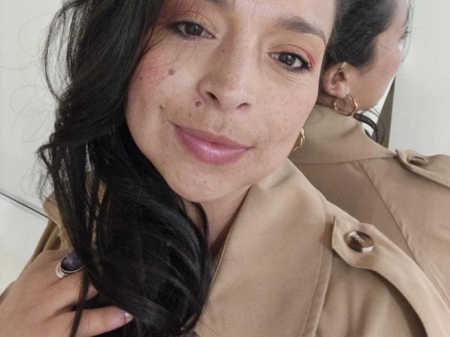 StangelarRivera with long black hair, brown eyes, and freckles, wearing a beige coat, smiling, holding her hair, and showing small tits in a mirror selfie.