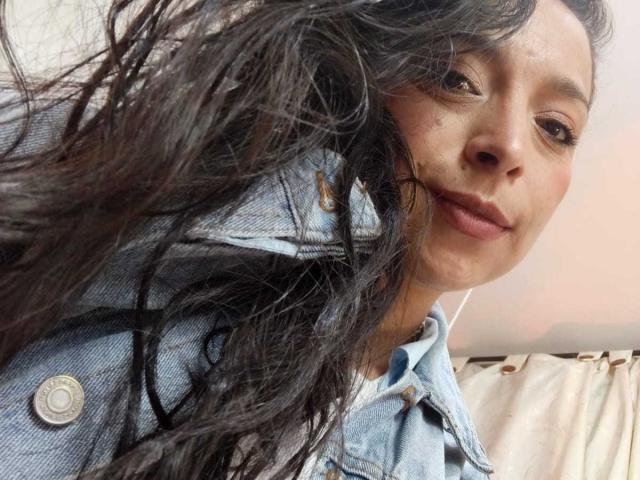 StangelarRivera with wavy black hair, light brown skin, and white earbuds, wearing a blue denim jacket, smiling slightly, showing her nose and lips.