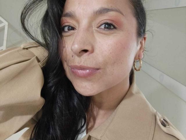 StangelarRivera has dark hair, brown eyes, and is wearing a beige uniform with gold hoop earrings. She's smiling with slightly freckled skin and pink lipstick.