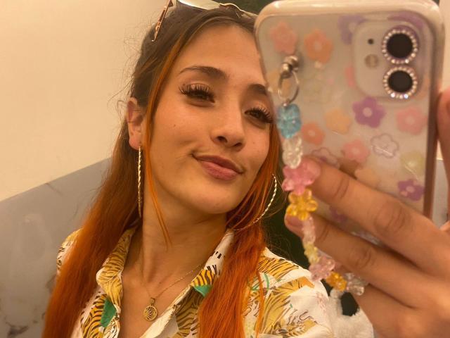 ZoeTaylorl with orange-dyed hair, wearing a floral shirt, hoop earrings, and a necklace, smiling at her phone, holding it up with colorful flower stickers.