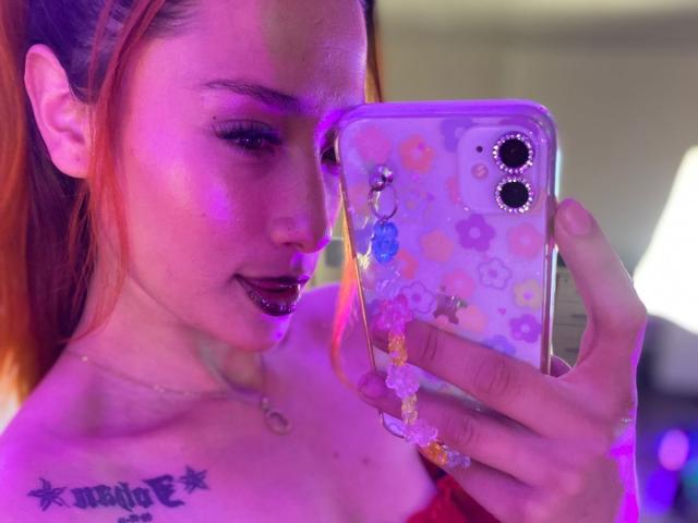 ZoeTaylorl with orange hair, dark lipstick, and tattoos, holding a phone with a floral case, looking at the screen with a slight smile, purple light in the background.