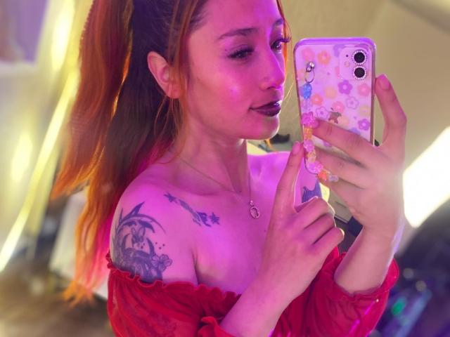 ZoeTaylorl with pink-orange hair in a ponytail, red off-shoulder top, black lipstick, holding a phone with a pink floral case, showing tattoos on her shoulder and chest.