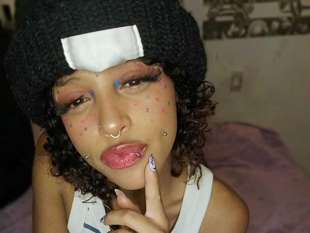 LunaKeller with curly black hair, black beanie, colorful face paint, nose ring, and lip piercing, looking at camera with finger on chin, wearing white tank top.