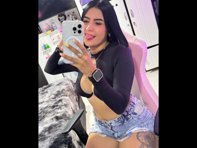 AmaraDeluxe with black hair, tongue out, wearing a black long-sleeve top, exposing her tits, and blue denim shorts, taking a selfie with a white iPhone in a room.