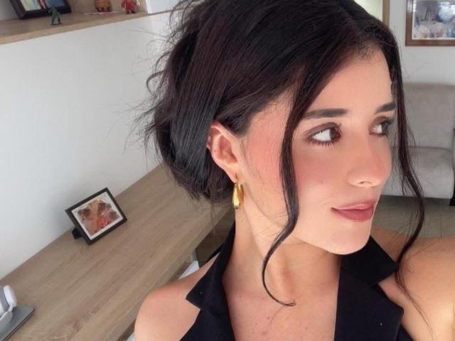VickyMegan with dark hair in a messy updo, wearing gold earrings, black sleeveless top, and subtle makeup, looking to the side with a slight smile.