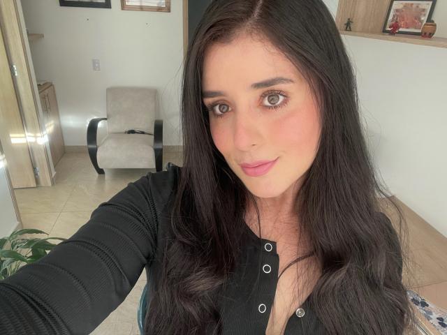 VickyMegan with long black hair, pink blush, and black buttoned top, showing small tits, smiling, green eyes, pink lipstick, and holding a phone for a selfie.