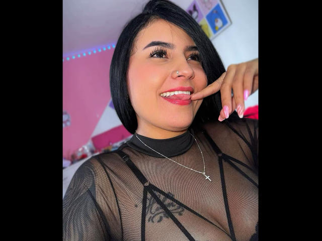 AmaraDeluxe with black hair, wearing sheer black top, showing tits, finger in mouth, pink nails, cross necklace, smiling, nose ring, medium skin, pink wall in background.