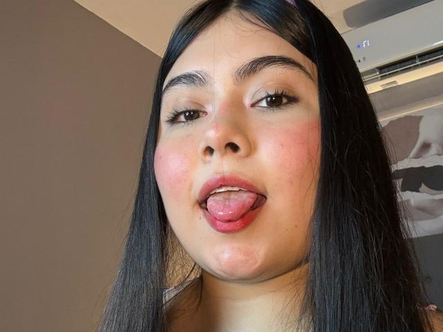 AriianaMills has long black hair, brown eyes, and is sticking out her tongue with her pink lips and slightly flushed cheeks. Her face is centered in the image.