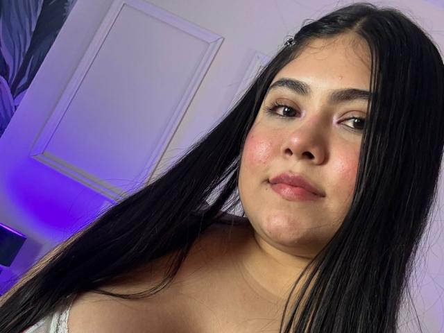 AriianaMills has long black hair, brown eyes, and a slight smile with pink cheeks. She's wearing a white top, showing her bare shoulders and a small pimple on her chin.