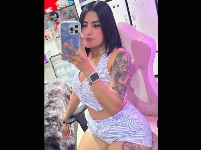AmaraDeluxe with black hair, wearing a blue striped crop top and shorts, holding a phone, showing a tattooed arm, sitting on a pink chair, looking at the camera.