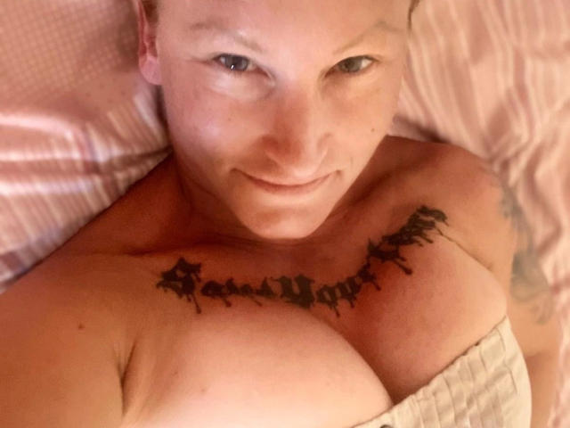 MelissaStrip-hot with a smile, looking up, has black tattoo across her chest, exposing her big tits, light skin, blue eyes, and a pink bedsheet in the background.