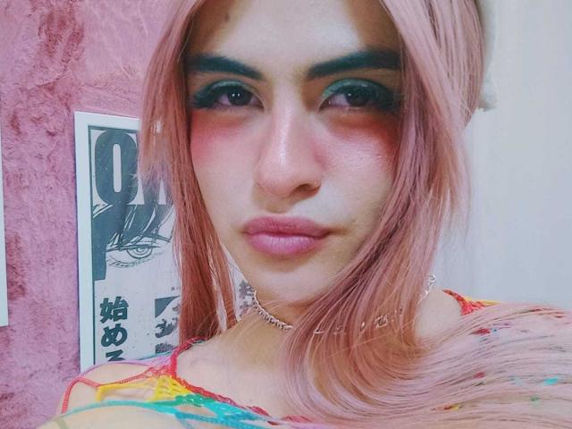 EmillyHall con i capelli rosa, il trucco occhi verde scuro, il blush rosa e le labbra rosa, indossa una maglia colorata, una collana e guarda la fotocamera con un'espressione seria..