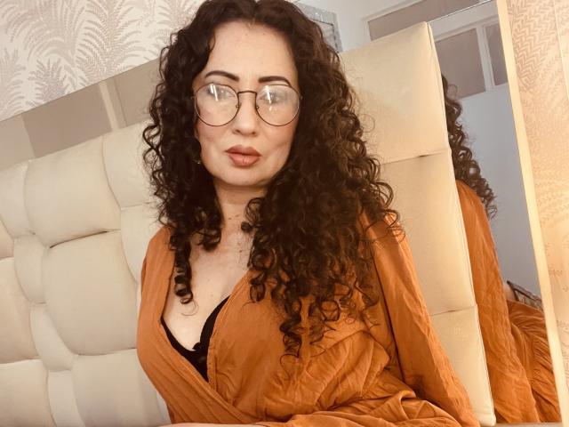 SamanthaAntolin with curly black hair, glasses, and orange shirt, showing her tits in a black bra, sitting on a beige tufted headboard, looking at the camera.