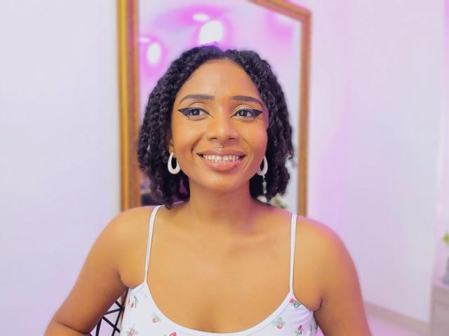 Megan Williams, con cabello negro rizado, sonriendo, vestida con un vestido blanco de tirantes finos con estampados de flores rosas, pequeños aretes y maquillaje con delineador de ojos llamativo. Sus senos son pequeños y su vulva no es visible..