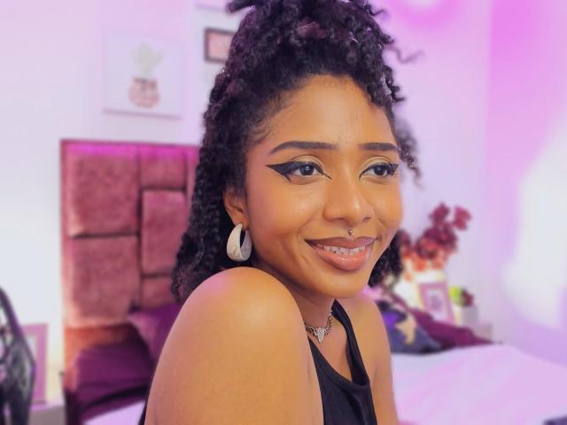 Megan Williams, con cabello negro rizado, piel oscura y delineador de ojos alado, sonriendo a la cámara, vestida con una blusa negra, aretes blancos y un collar, mostrando su atractivo trasero..