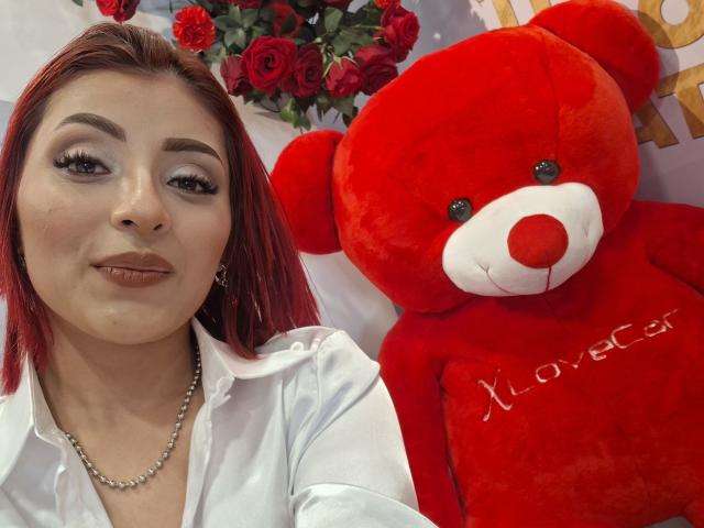 LizBonnet, con cabello rojo, camisa blanca y collar de perlas, sosteniendo un osito de peluche rojo con una nariz roja y la inscripción "Love Cat" en su pecho. Sus senos son parcialmente visibles..