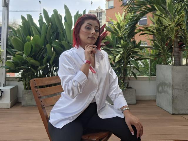LizBonnet, con cabello rojo, camisa blanca, pantalones negros, sentada en una silla de madera, sosteniendo un teléfono en la oreja, con una pulsera rosa, con una expresión segura, de busto medio, en una azotea con plantas..