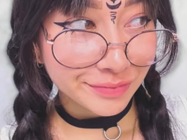 Genesiss with dark brown hair in braids, wearing round glasses, black choker, and pink lipstick, has tattoos above eyebrows, smiling with pinkish skin, and a cute face.