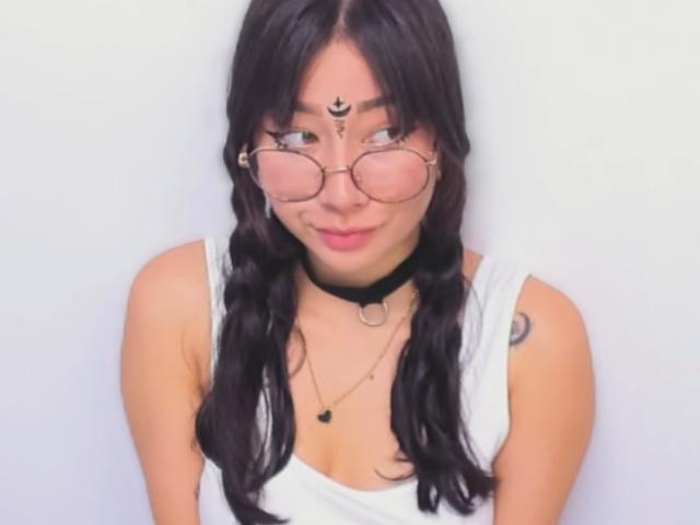 Genesiss has black hair in pigtails, wearing round glasses, a white tank top, black choker, and necklace. She's smiling with a tattoo on her right shoulder.