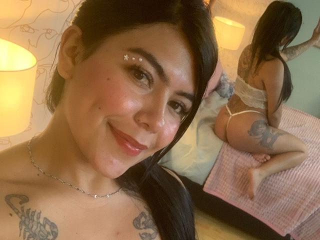 CatalinaReiyna with dark hair, smiling, wearing a white thong, showing tattoos on her chest and thigh, and her ass in a mirror reflection. She's topless with small breasts.
