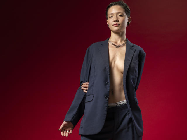 LissyLuxe, shirtless in a dark blue suit, hands behind back, necklace, short brown hair, slightly open jacket, visible abs, Calvin Klein underwear, standing against red background.