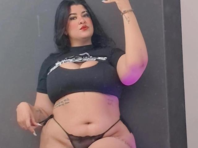 RashaelBig with black hair, red lipstick, and tattoos, wearing a black crop top and black thong, showing her big tits and big ass, holding her thong.