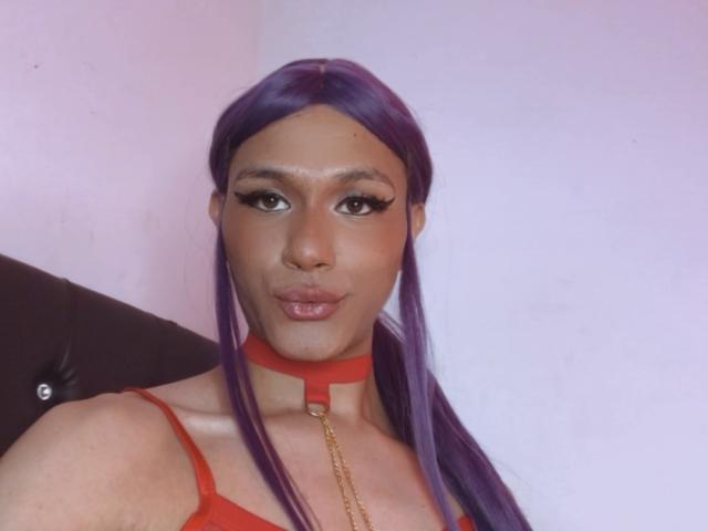 Holyshweet69 with purple hair, wearing a red choker and red top, has a sexy face, full lips, and a chain hanging from the choker, with a cute, confident expression.