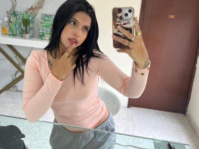 NyraVanel con capelli neri, maglia a maniche lunghe rosa, pantaloni grigi, un dito sul mento, che tiene un telefono, riflesso nello specchio, anello dorato, sedia bianca, tavolo di vetro, porta marrone..
