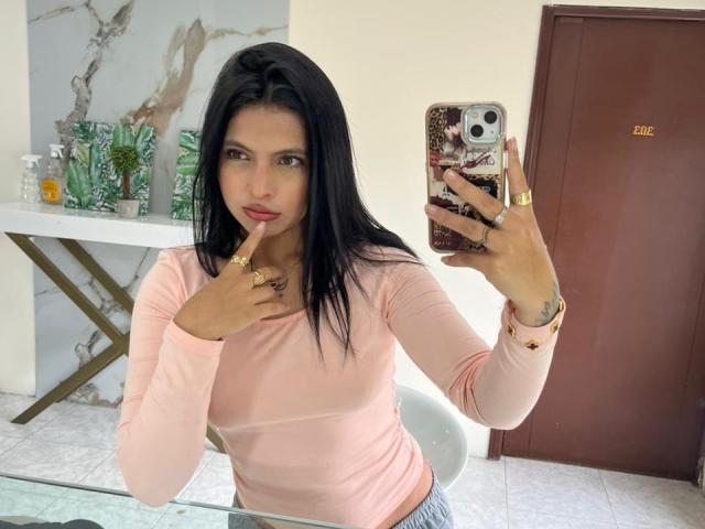 NyraVanel with black hair, pink long-sleeve shirt, finger on lips, holding phone, gold rings, looking at mirror, marble wall, brown door, glass table, gray pants.
