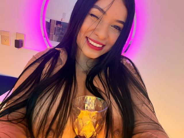 AliceNova with long black hair, smiling, holding a glass with ice, pink neon ring light behind her, visible cleavage, pink lipstick, white wall, and electrical outlet on the left.