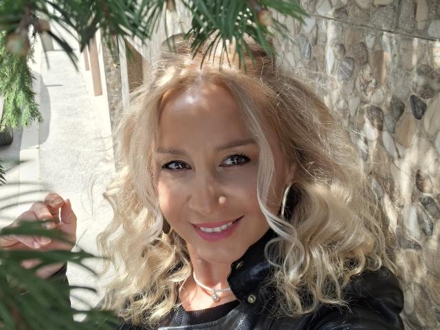 Merlynon has blonde curly hair, tan skin, and is smiling with pink lipstick. She's wearing a black jacket and has a necklace. Pine leaves are in the foreground.