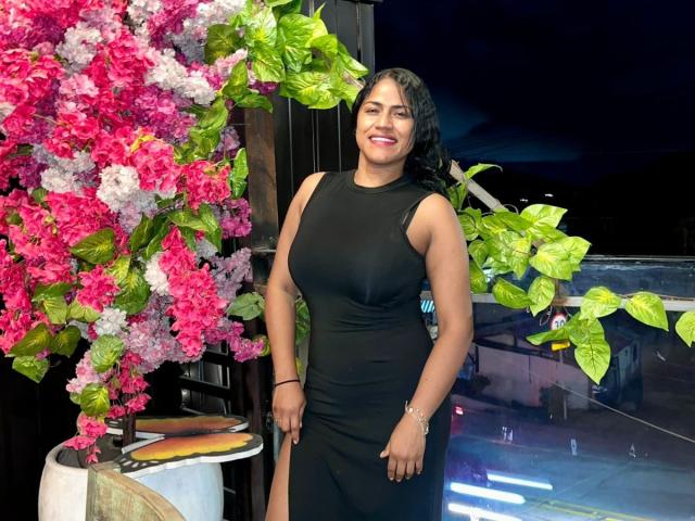 IvannaDuran with curvy, dark-skinned ass in a tight black dress, smiling at pink and purple flowers with green leaves. Her black hair is wavy, and she's wearing a bracelet.
