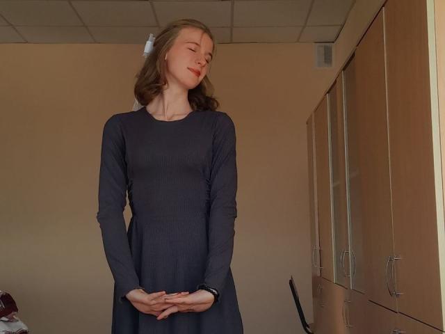DebbyPennimpede, with eyes closed and hands folded, stands in a room wearing a long, dark dress, her ass subtly visible, blonde hair, white hairpin, wooden cabinets on the right.