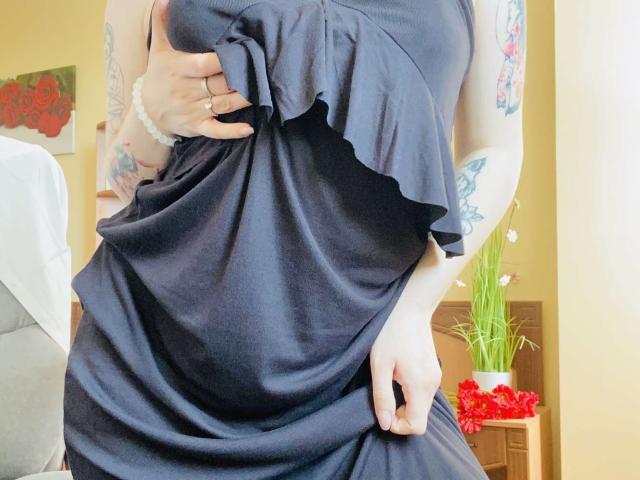 KarKutie's hand holds up a ruffled, dark blue dress, revealing her tits and tattoos on her right arm. She's wearing a white bracelet, and her nails are painted.