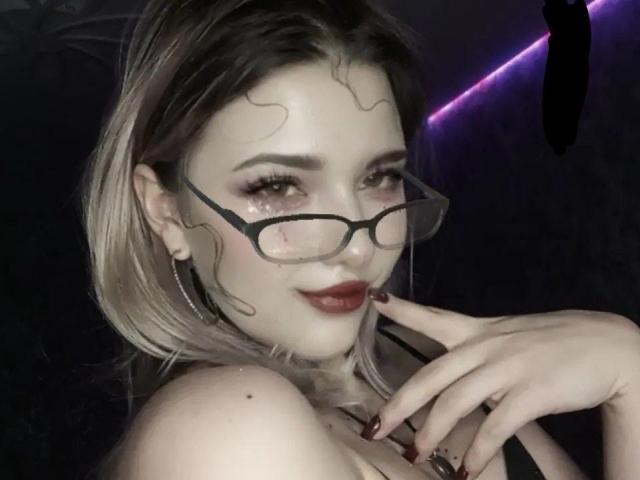 Adriane Pittillo, con cabello rubio ondulado, gafas y lápiz labial rojo oscuro, tocándose la barbilla con la mano, mostrando sus senos. Sus uñas están pintadas de rojo oscuro..