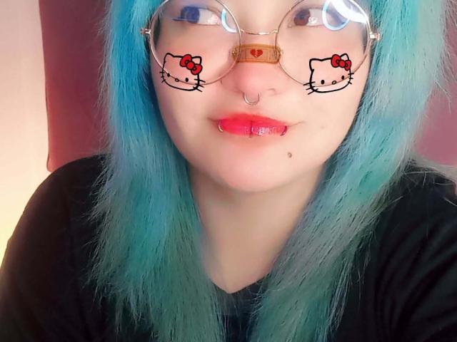 LuckyLeahh with blue hair, glasses featuring Hello Kitty stickers, nose ring, lip ring, red lipstick, black shirt, and a small mole on her chin.