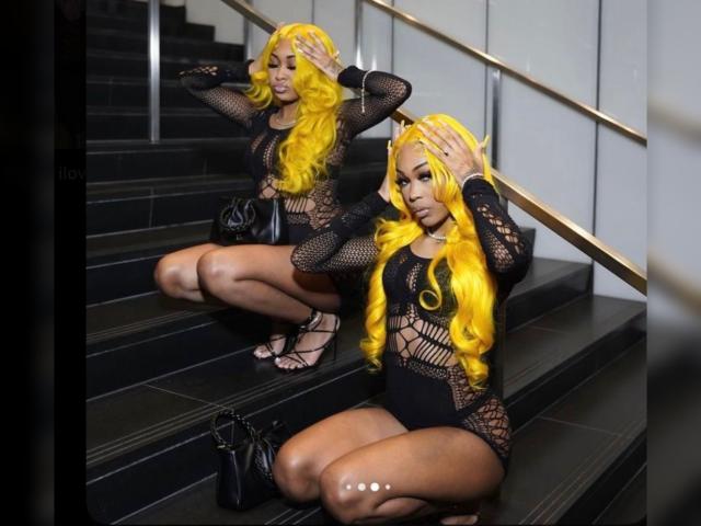 CoochieFlames tiene el pelo largo y rubio, un body de malla negra y tacones negros, está agachada en unas escaleras oscuras, con las manos en el pelo, luciendo sexy, y tiene un bolso negro en el suelo..
