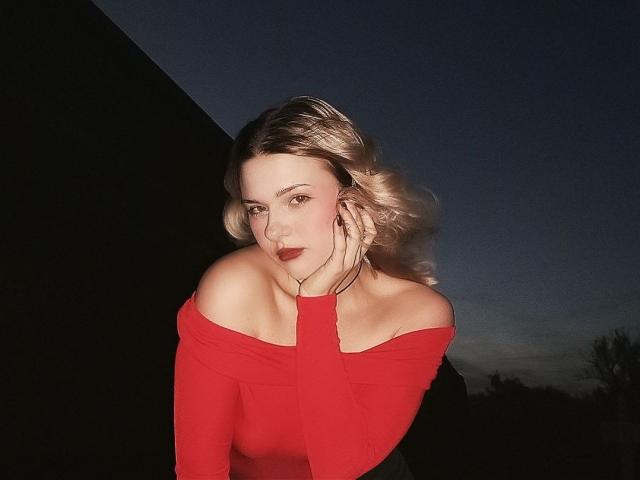 Adriane Pittillo, com cabelo loiro, blusa vermelha com um ombro à mostra, mão no rosto, batom escuro, olhando para a câmera, fundo de céu noturno, seios visíveis, sorriso discreto, brincos..