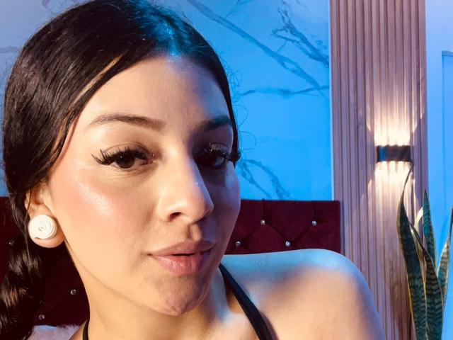 LonxEva has dark hair, light skin, and is wearing white earbuds with thick eyelashes, glossy pink lips, and a black halter top. Her ass is partially visible.