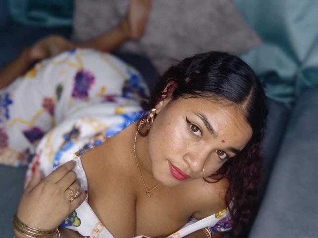 DarsshaHara, con i capelli scuri e ricci, gli occhi marroni e il rossetto rosso, indossa un abito floreale, mostrando il suo seno prosperoso, è sdraiata su un divano e guarda verso l'alto con un leggero sorriso..