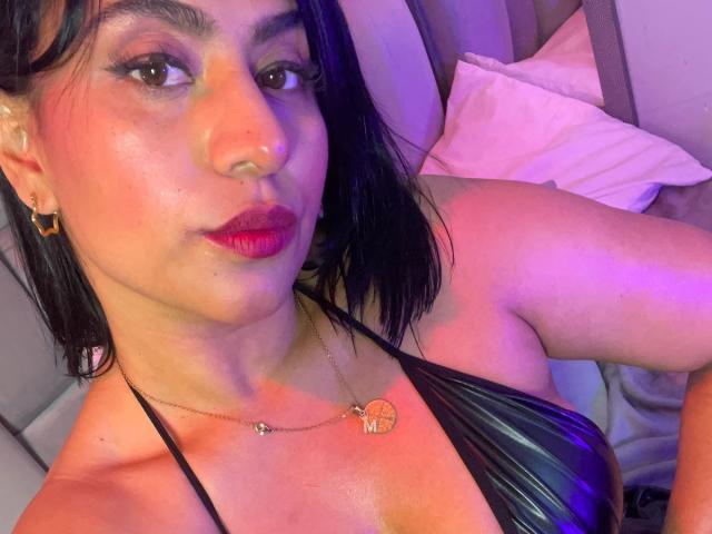 DiannaRosse with dark hair, red lipstick, shiny skin, small nose ring, gold earrings, black halter top, heart necklace, purple-blue light, looking at camera, showing her tits.