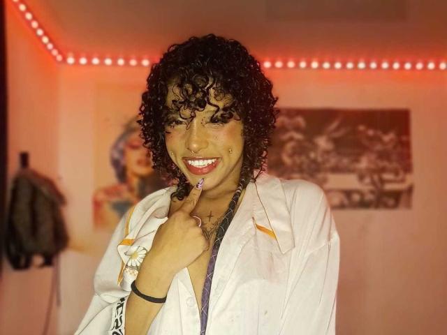LunaKeller with curly black hair, smiling, wearing a white jacket, holding her finger to her red lipstick, has a nose ring and a bracelet, in a room with red LED lights.
