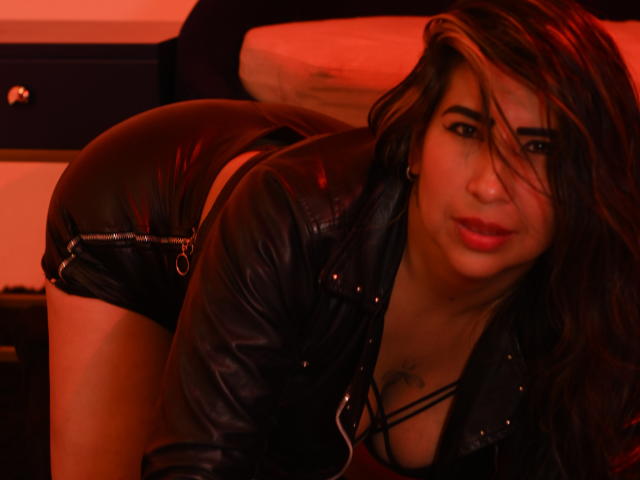 SahamaranDesiri with long brown hair, red lipstick, black leather jacket, and tight black leather shorts, bent over, showing her ass, in a red-lit room. She's looking at the camera.