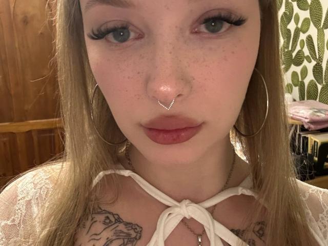 Rossana Semone tem sardas, olhos azuis e um piercing no nariz. Ela está usando brincos de argola, uma blusa branca de renda e tem tatuagens no peito. Seus lábios são levemente rosados..