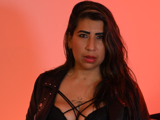 SahamaranDesiri with dark hair, red lips, black strappy top, and black leather jacket, showing small tattoo below tits, against red background. She looks slightly annoyed.