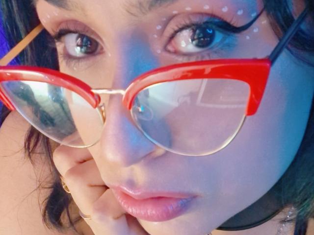 RoxanneHan with glossy red cat-eye glasses, pale skin, and dark brown hair, looking into the camera, hand near her mouth, wearing pink lipstick and a black choker.