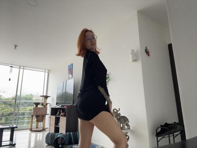 SarahRousen with red hair, glasses, black dress, and exposed ass, standing in a bright room, looking back, showing tattoo on right arm, and a cute cat on the floor.